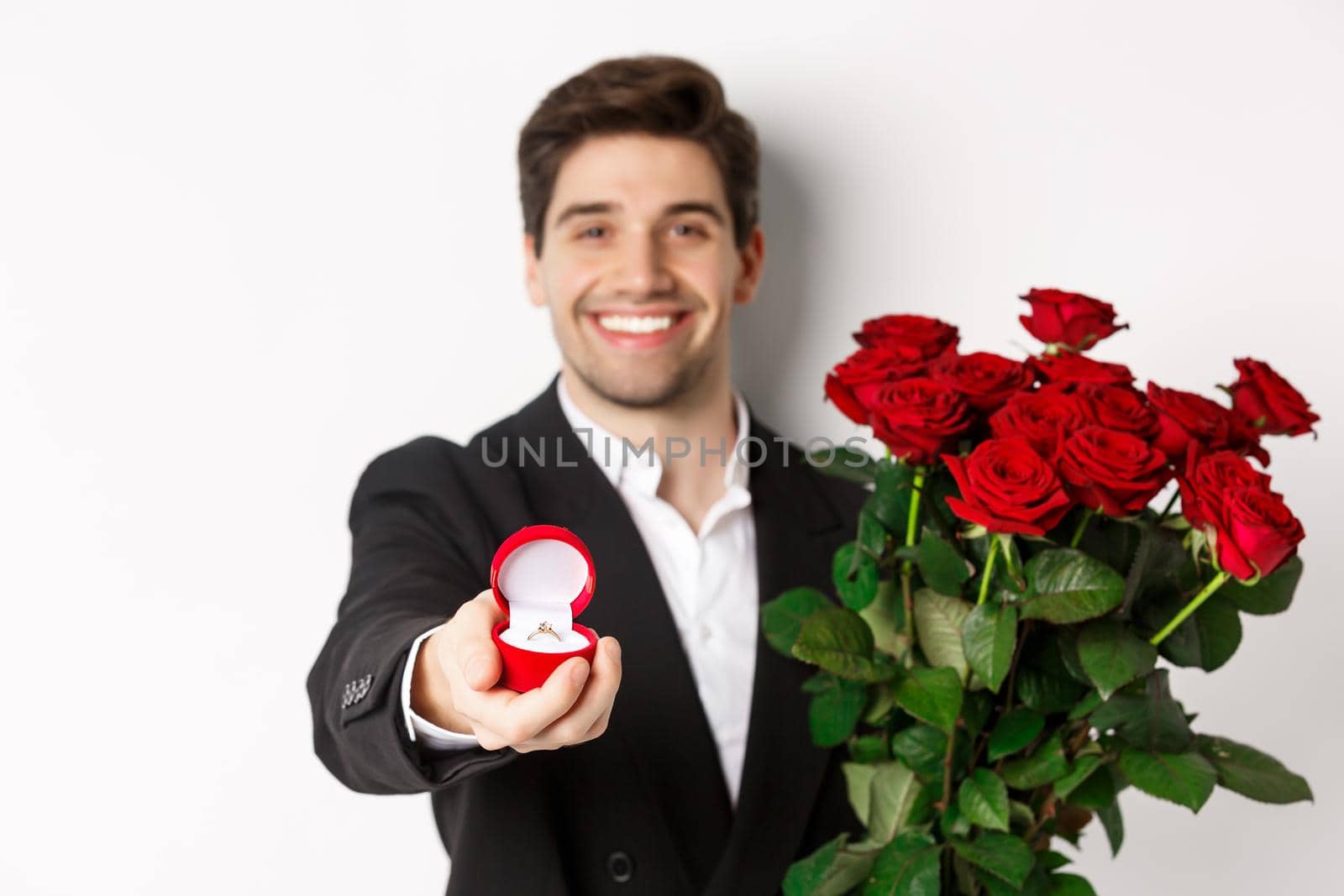 Close-up of attractive man in suit, holding bouquet of roses giving an engagement ring, proposing to girlfriend, standing against white background by Benzoix