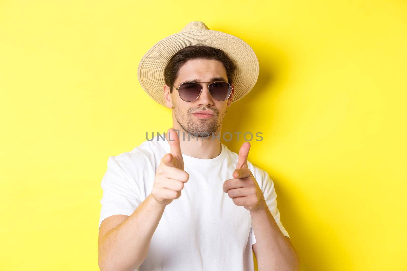 Concept of tourism and vacation. Close-up of cool guy in summer hat and sunglasses pointing finger guns at camera, standing over yellow background.