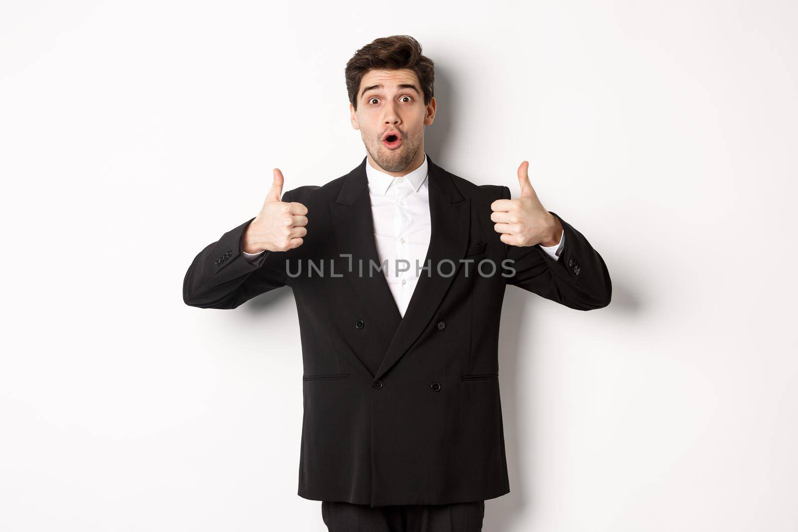 Concept of new year party, celebration and lifestyle. Portrait of excited handsome businessman in formal suit, showing thumbs-up in approval, recommending shop, standing over white background by Benzoix