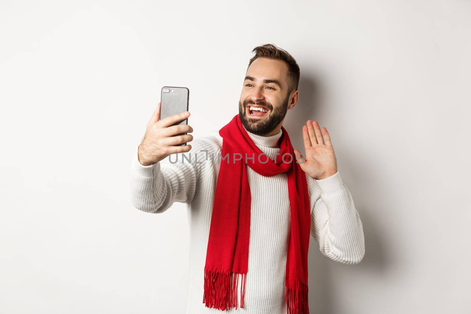 Handsome bearded man video calling friends, saying hello and waving hand at mobile phone, standing in sweater and red scarf against white background.