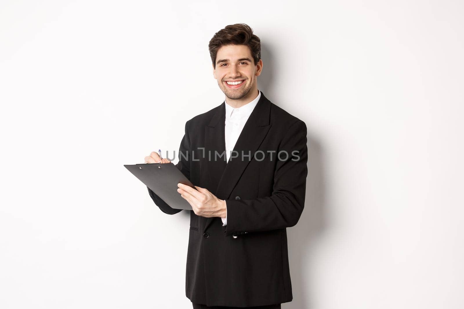Portrait of confident businessman in black suit, signing documents and smiling, standing happy against white background.