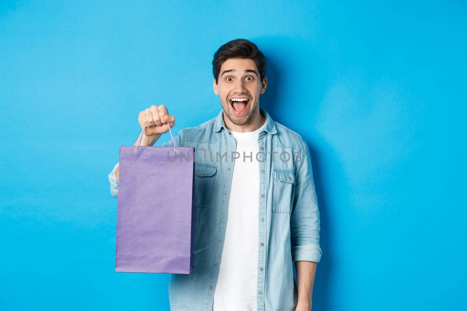 Concept of shopping, holidays and lifestyle. Excited handsome guy holding paper bag with gift and looking happy, standing over blue background.