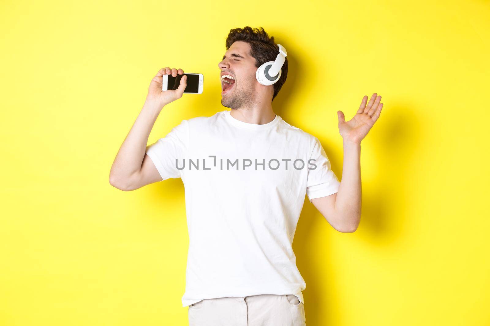 Happy guy playing karaoke app in headphones, singing into smartphone microphone, standing over yellow background.