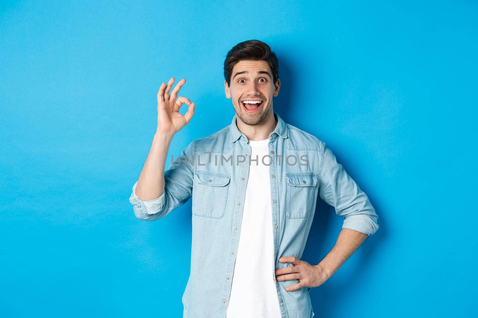 Smiling happy man showing ok sign and looking pleased, approving something good, standing against blue background by Benzoix