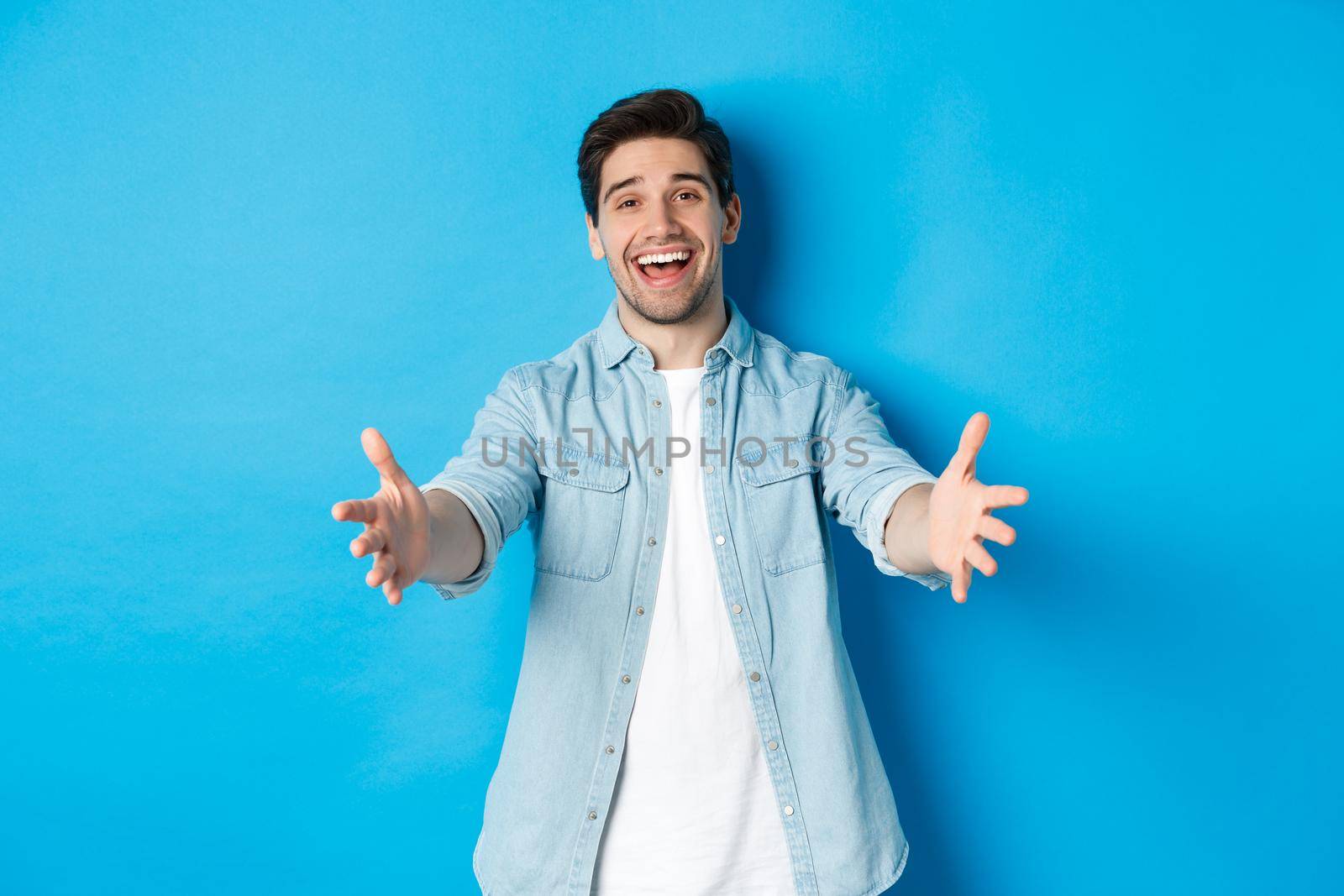 Friendly man stretching hands forward and welcome you, greeting guests or reaching for hugs, standing against blue background.