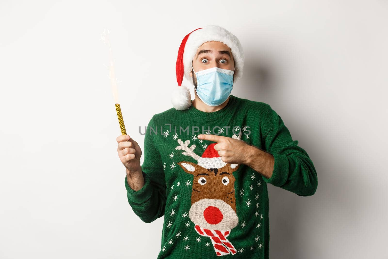 Concept of covid-19 and Christmas holidays. Excited young man in face mask, pointing finger at party sparkler, celebrating New Year during quarantine, standing over white background.