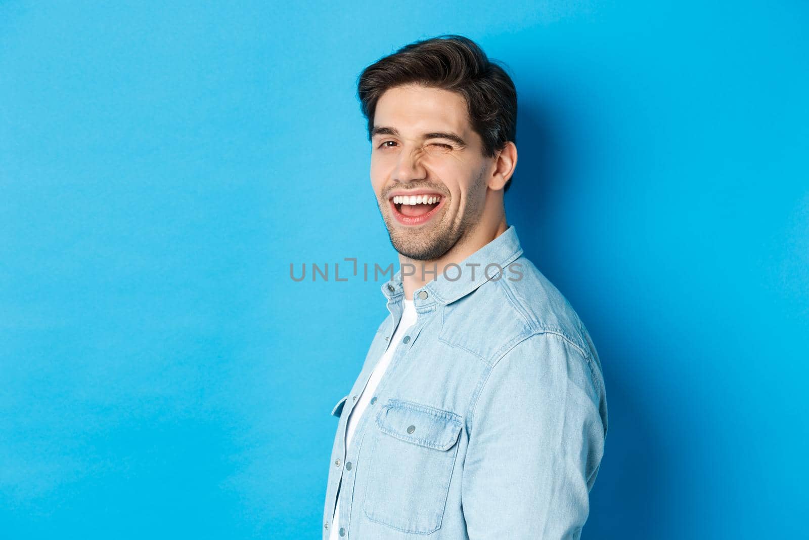 Happy and confident man turn head at camera, winking and smiling, standing over blue background.