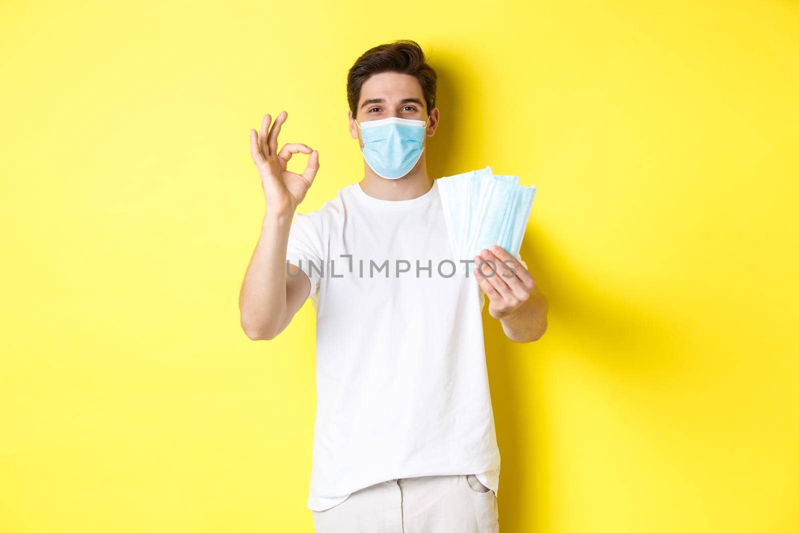 Concept of covid-19, quarantine and preventive measures. Satisfied man showing okay sign and giving medical masks, standing over yellow background.
