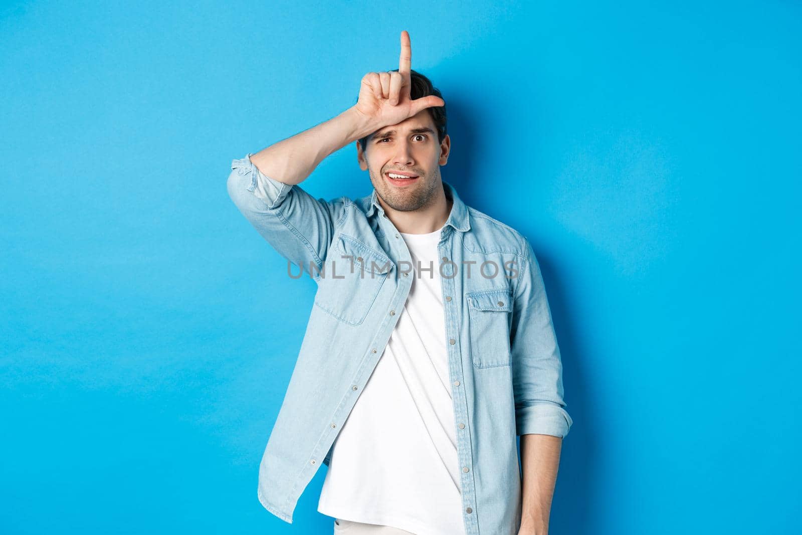 Rude guy mocking person for losing, showing loser sign on forehead and looking with dismay, standing against blue background.