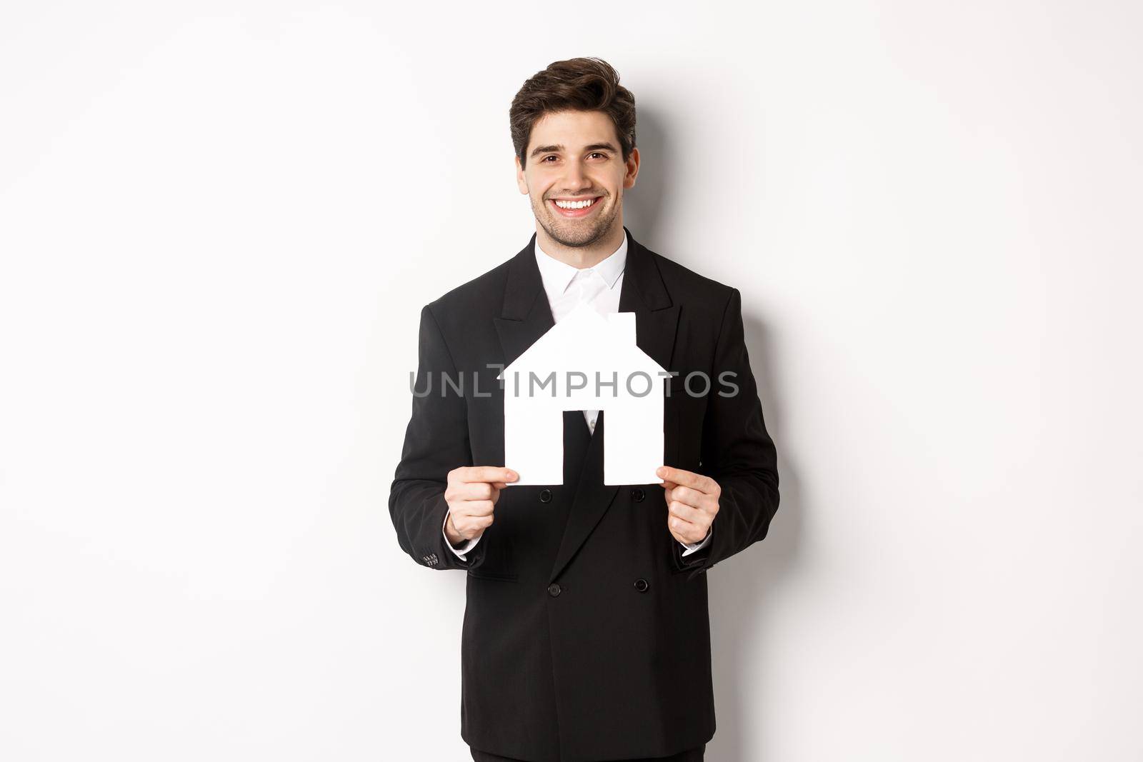 Image of handsome real estate agent in black suit showing home maket, smiling at camera, standing against white background.