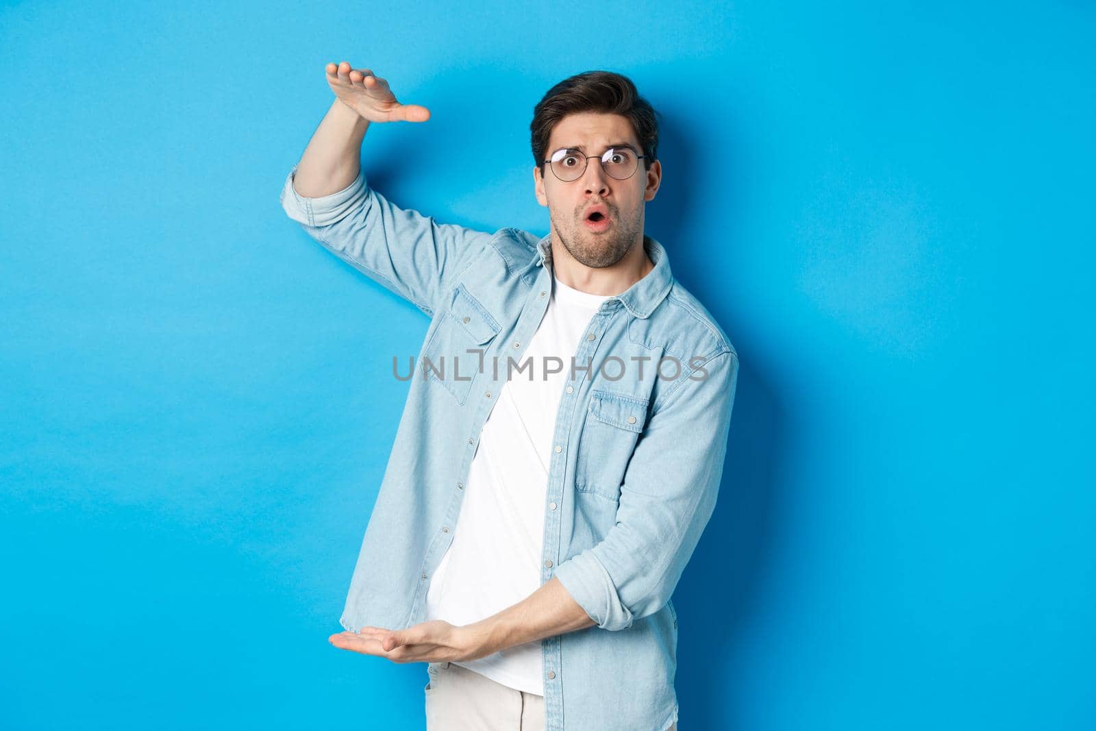 Confused handsome man showing big size, shaping large box and looking amazed, standing over blue background by Benzoix