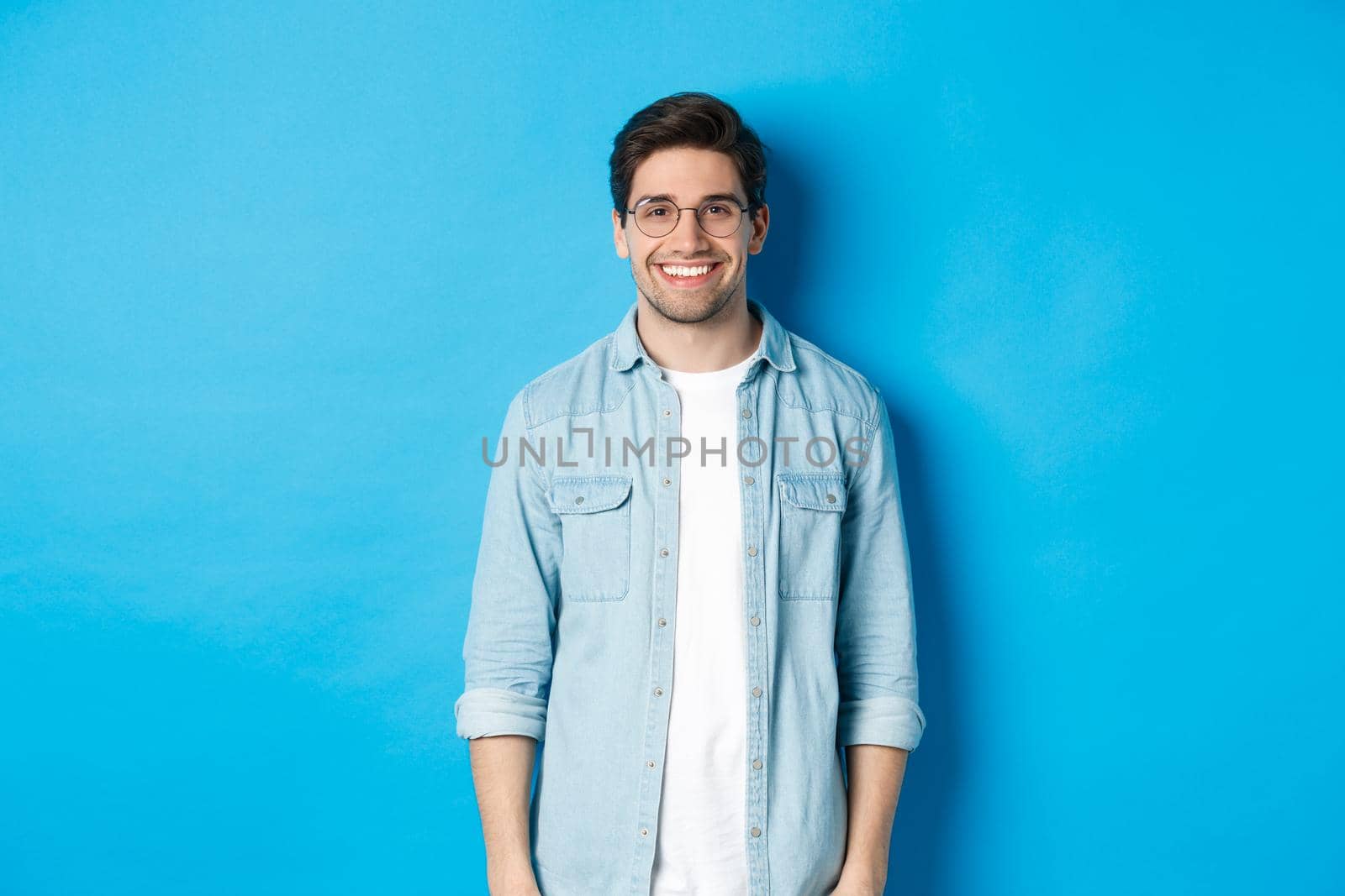 Young modern man in glasses and casual outfit standing against blue background, smiling happy at camera.