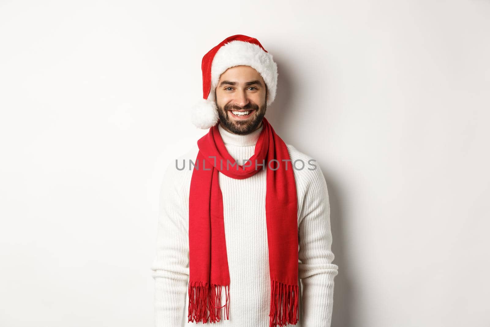 Christmas holidays. Smiling bearded man in santa hat and red scarf, smiling happy, standing against white background.