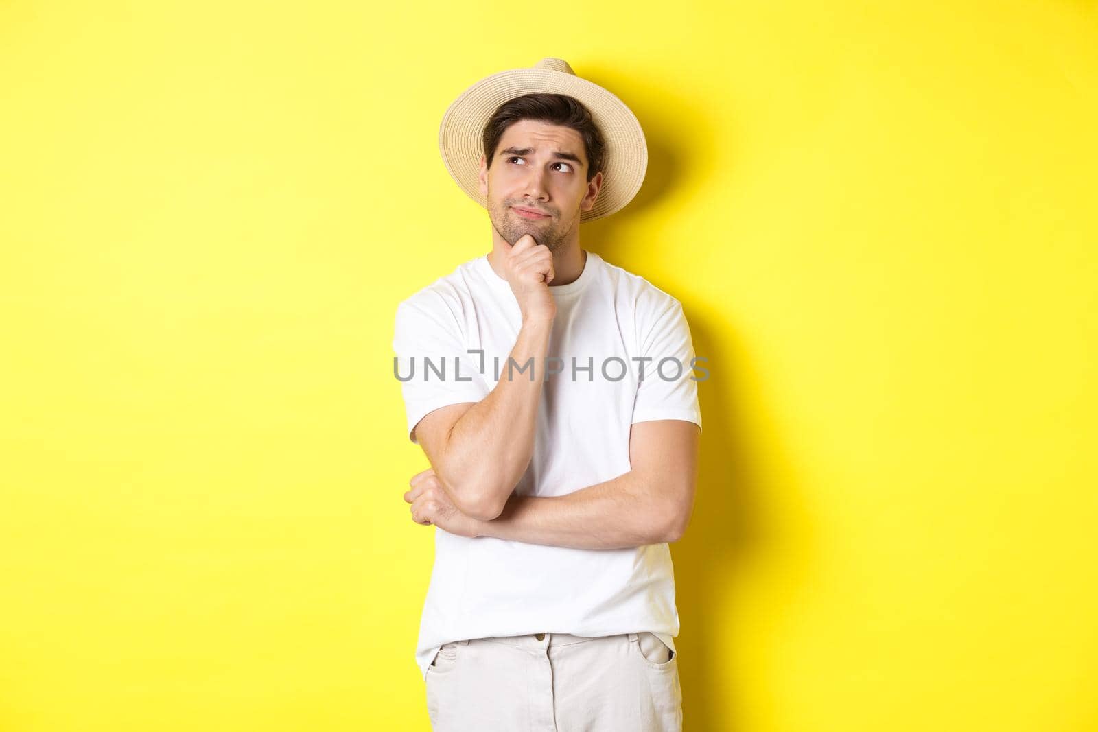 Concept of tourism and summer. Thoughtful man tourist pondering, looking at upper left corner and thinking, standing in straw hat and white t-shirt against yellow background.