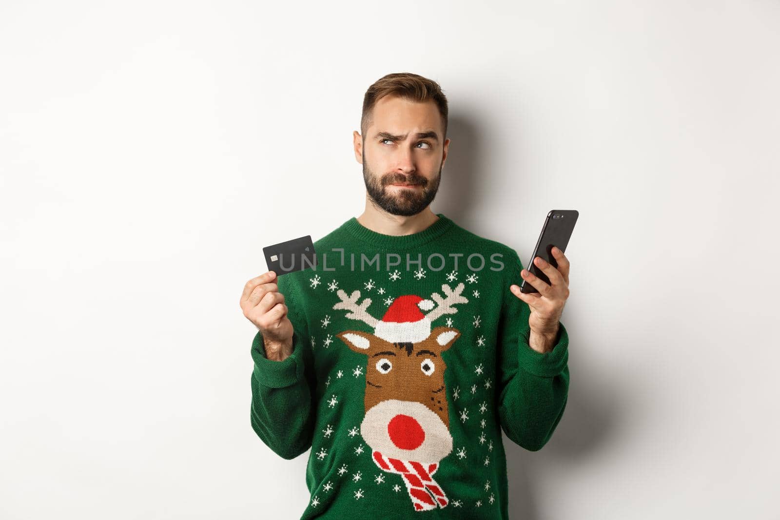 New Year, online shopping and christmas concept. Thoughtful man using mobile phone and credit card, thinking about gifts, standing over white background.
