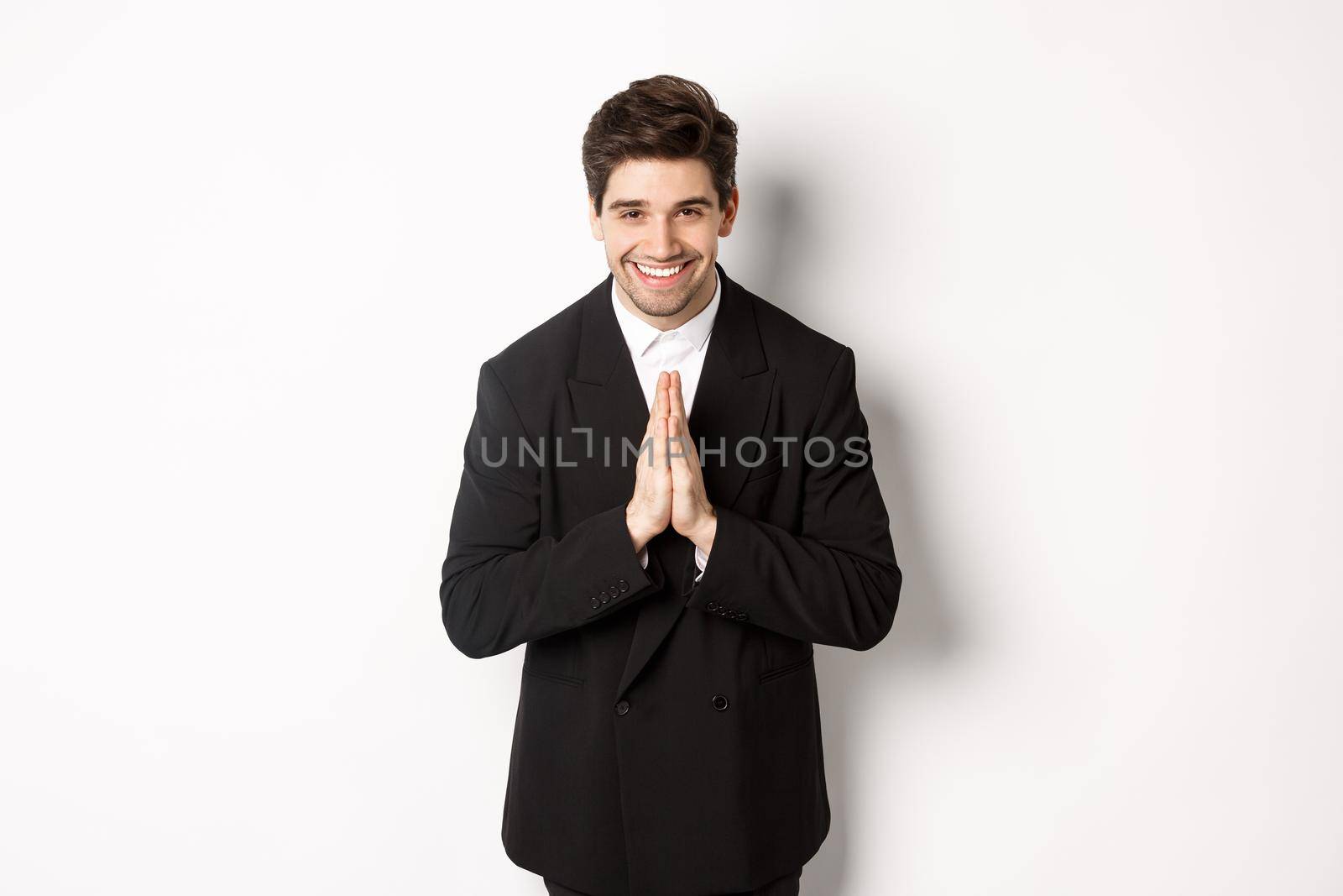 Portrait of handsome man in black suit, being grateful, saying thank you and bowing politely, smiling as holding hands together, expressing gratitude, standing over white background.