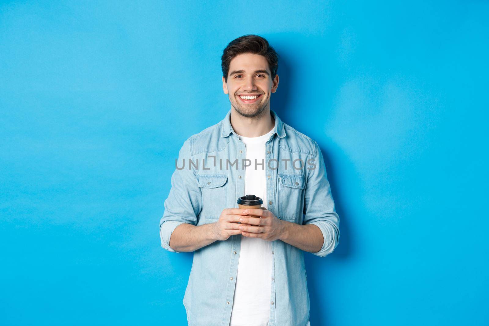 Young happy man drinking coffee from cafe takeaway, smiling pleased, standing against blue background.