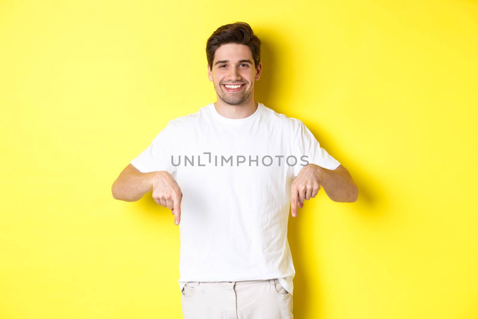 Attractive bearded man pointing fingers down, showing event banner, standing over yellow background.