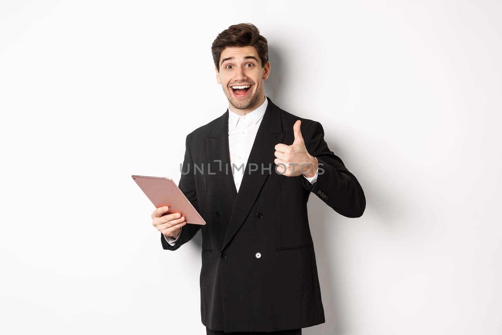 Portrait of handsome businessman in suit, showing thumb-up and making a compliment, praising something on digital tablet, standing over white background.