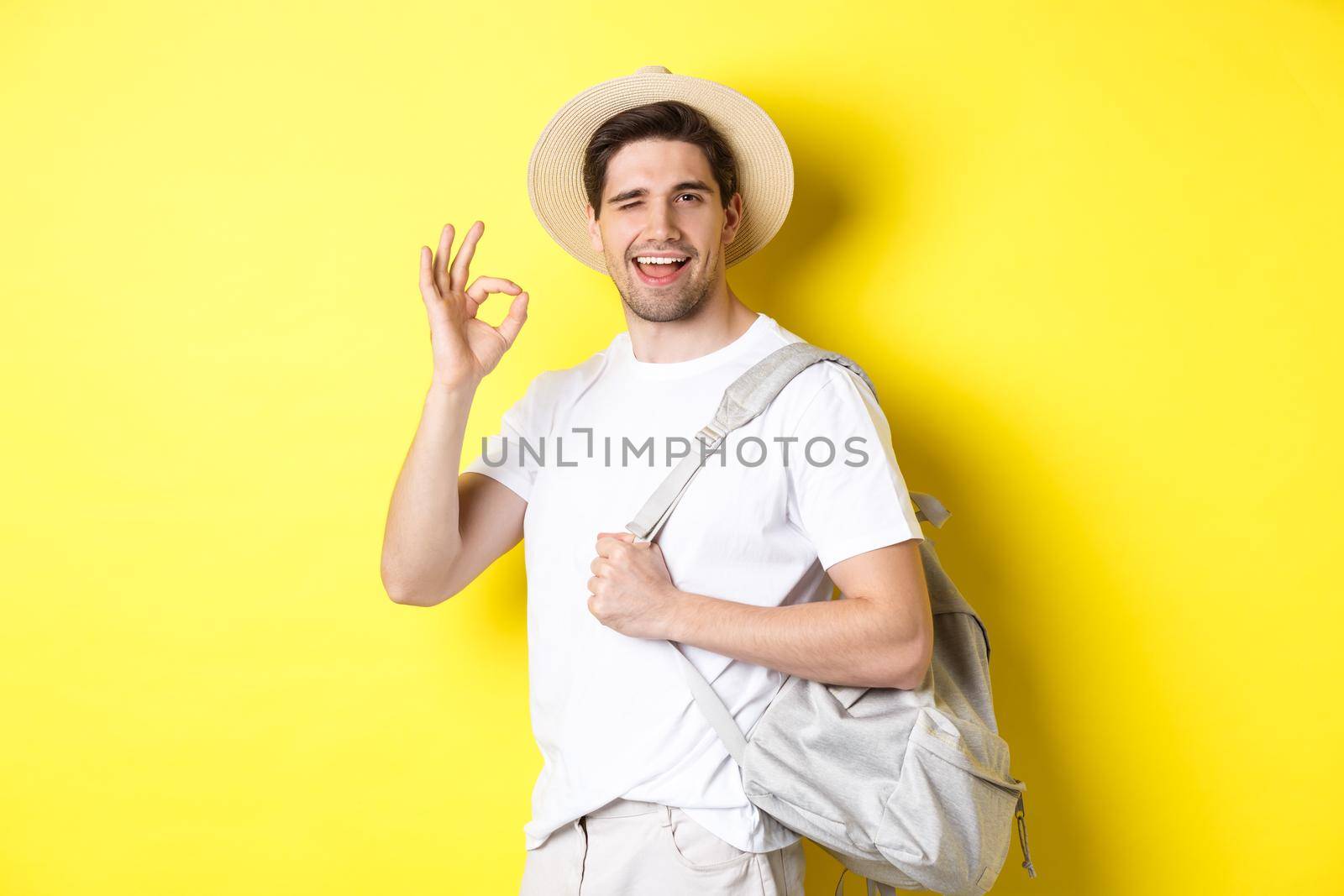 Tourism, travelling and holidays concept. Happy tourist going on vacation, holding backpack and showing okay sign smiling satisfied, standing against yellow background by Benzoix