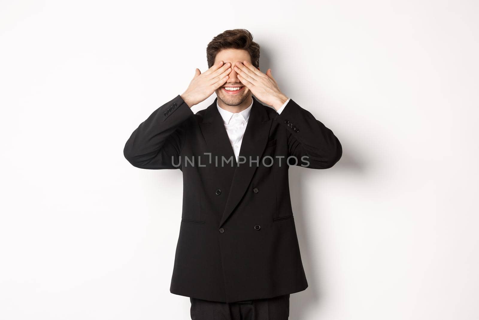 Image of handsome stylish man in black suit, waiting for christmas surprise, covering eyes with hands and smiling, anticipating presents, standing over white background by Benzoix