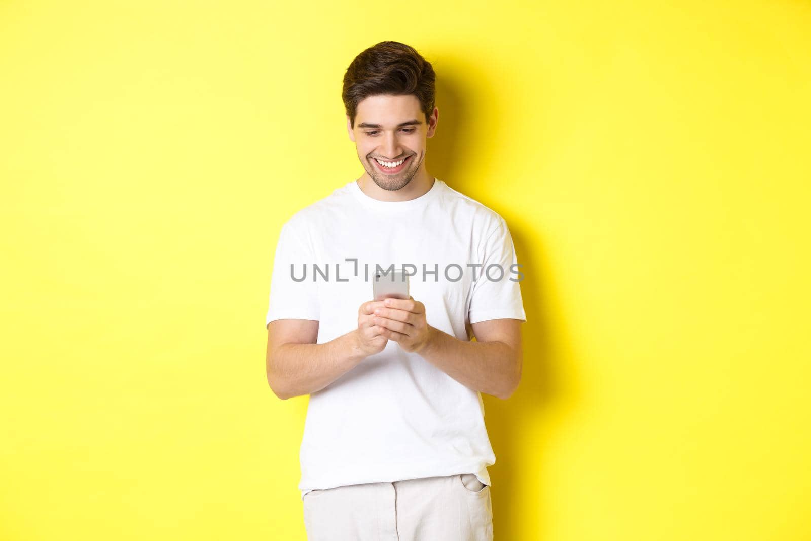 Young man reading text message on smartphone, looking at mobile phone screen and smiling, standing in white t-shirt against yellow background.