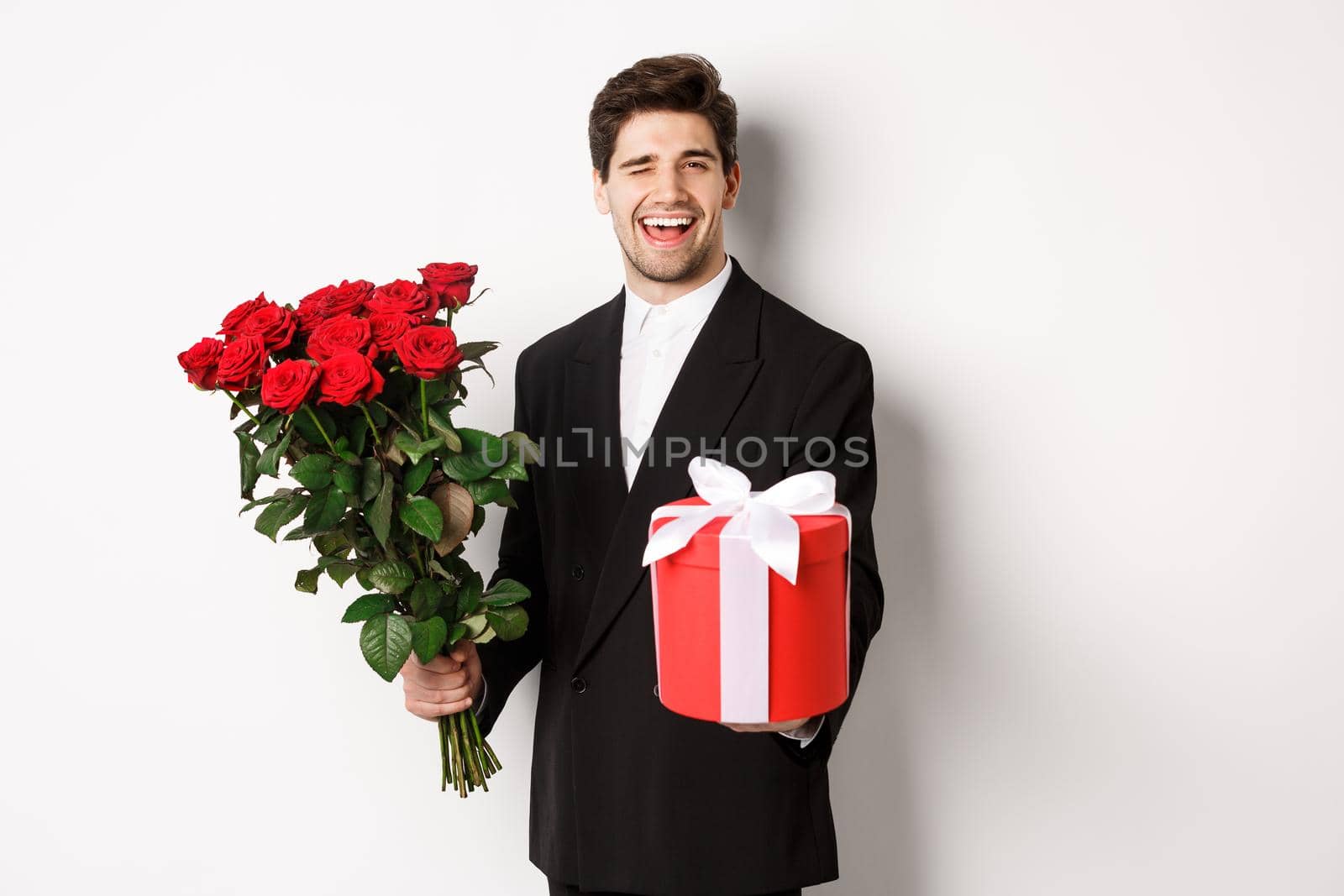 Concept of holidays, relationship and celebration. Charming young man in black suit, holding gift box and bouquet of roses, winking and smiling, standing against white background by Benzoix