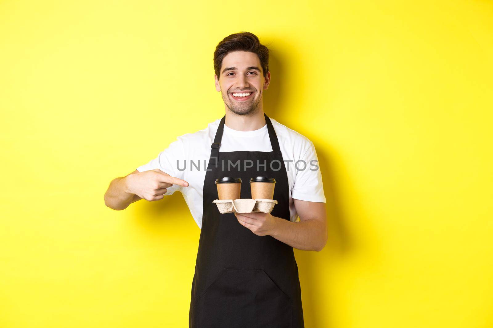 Handsome barista holding two cups of takeaway coffee, pointing finger at drinks and smiling, standing in black apron against yellow background by Benzoix