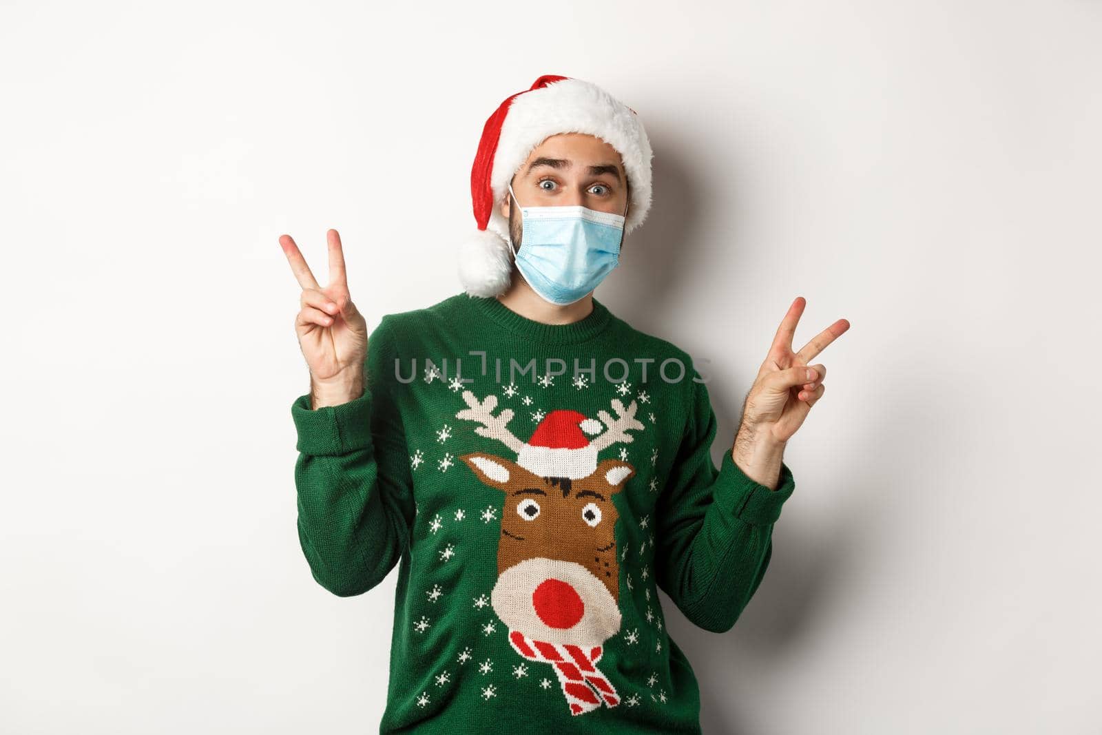 Concept of covid-19 and Christmas holidays. Happy man in medical mask and Santa hat showing peace signs, standing over white background.