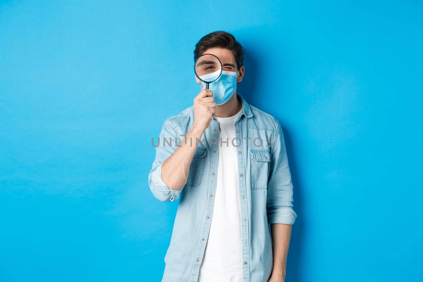 Concept of covid-19, social distancing and quarantine. Young man in medical mask searching for something, looking through magnifying glass, standing against blue background by Benzoix