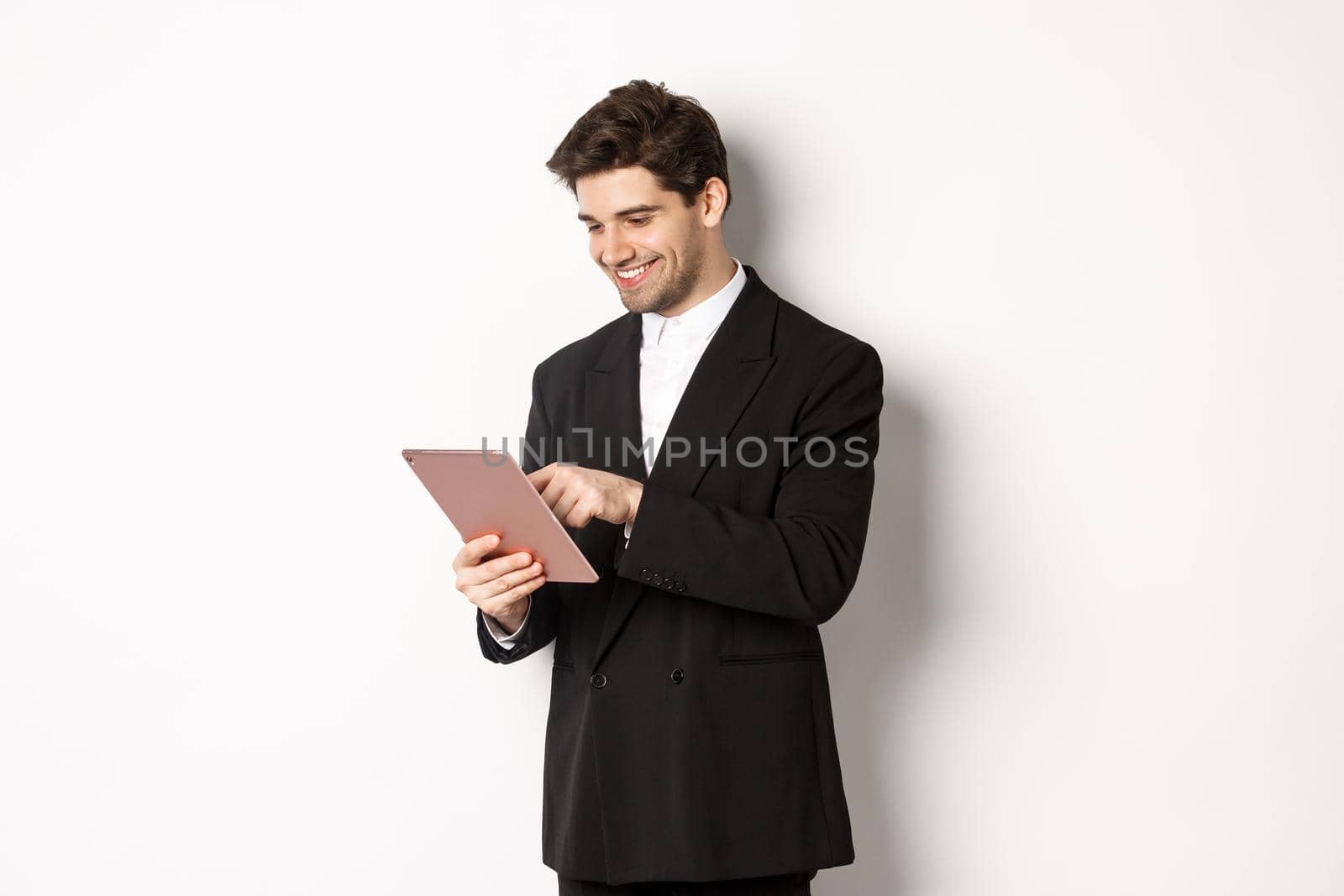 Portrait of attractive man in trendy suit, looking at digital tablet and smiling, shopping online, standing over white background.