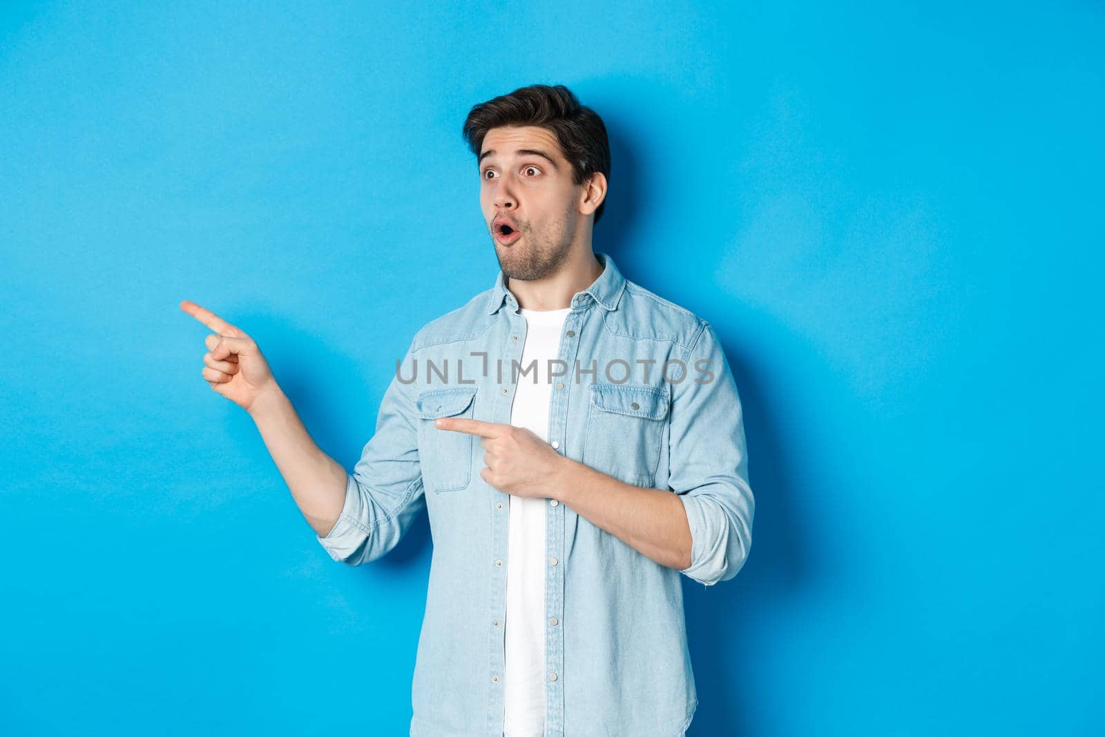 Surprised and amazed man looking at promotion, pointing fingers left at advertisement, standing over blue background by Benzoix
