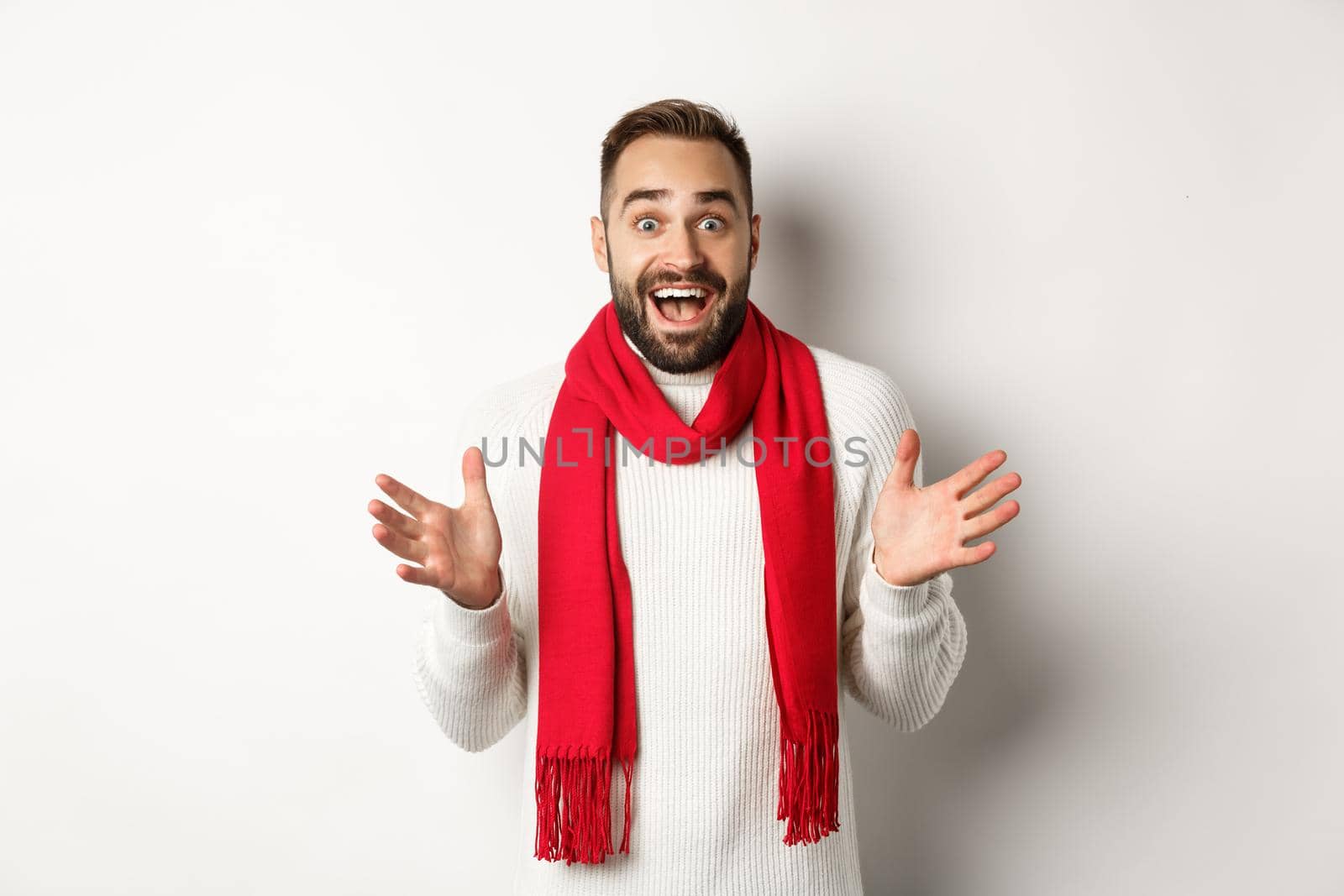 Christmas holidays and celebration concept. Bearded guy looking surprised at new year promo offers, gasping amazed, wearing red scarf and sweater, white background.