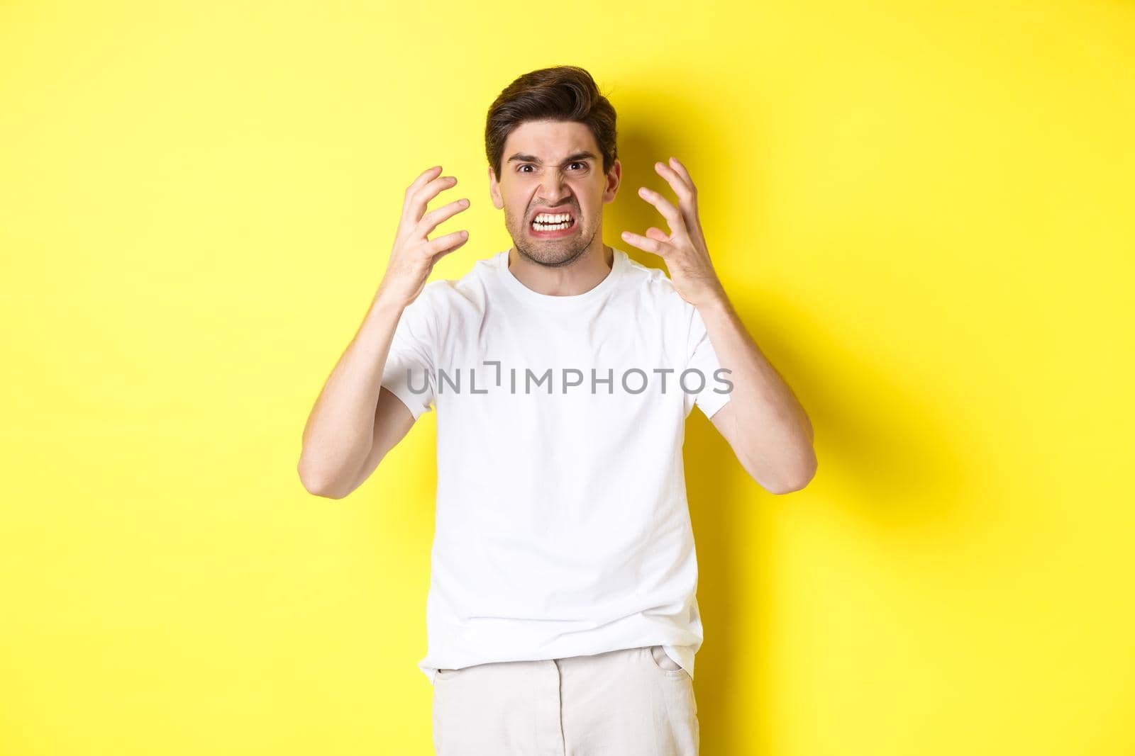 Angry man looking mad, grimacing and shaking hands furious, standing outraged against yellow background by Benzoix