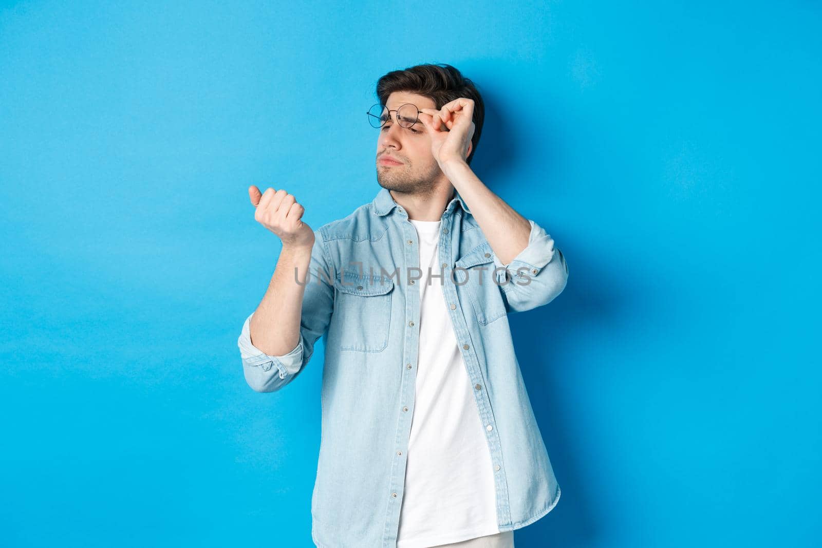 Young handsome man in glasses looking at his fingernails, checking manicure, standing over blue background by Benzoix