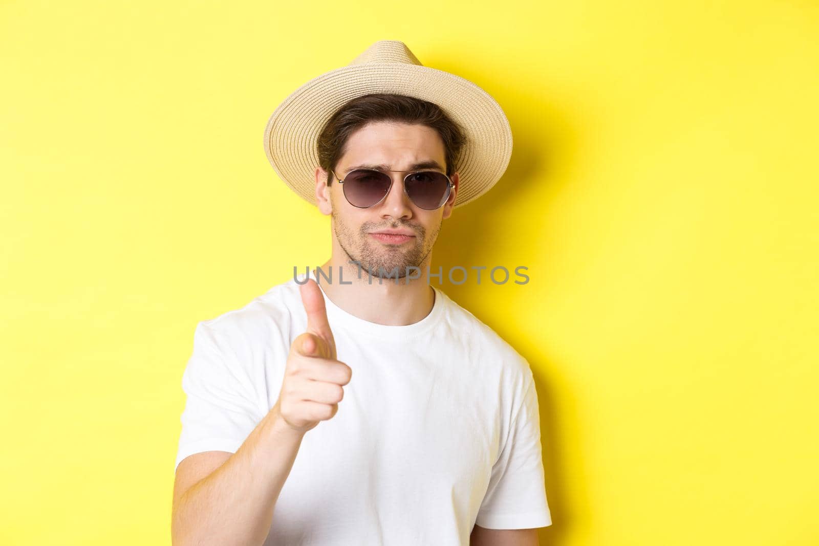 Concept of tourism and vacation. Close-up of cool guy in summer hat and sunglasses pointing finger at camera, standing over yellow background.