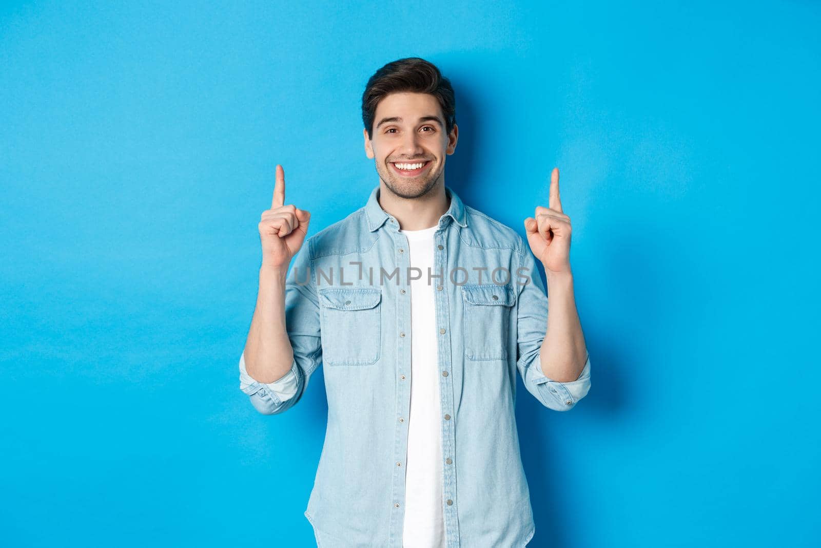 Portrait of handsome bearded man in casual outfit, smiling happy and pointing fingers up at copy space, standing over blue background.