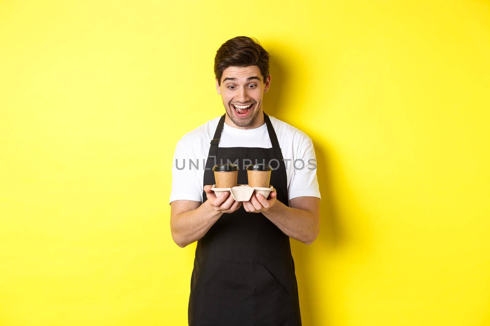 Waiter looking excited at two cups of takeaway coffee, wearing black apron, standing over yellow background by Benzoix