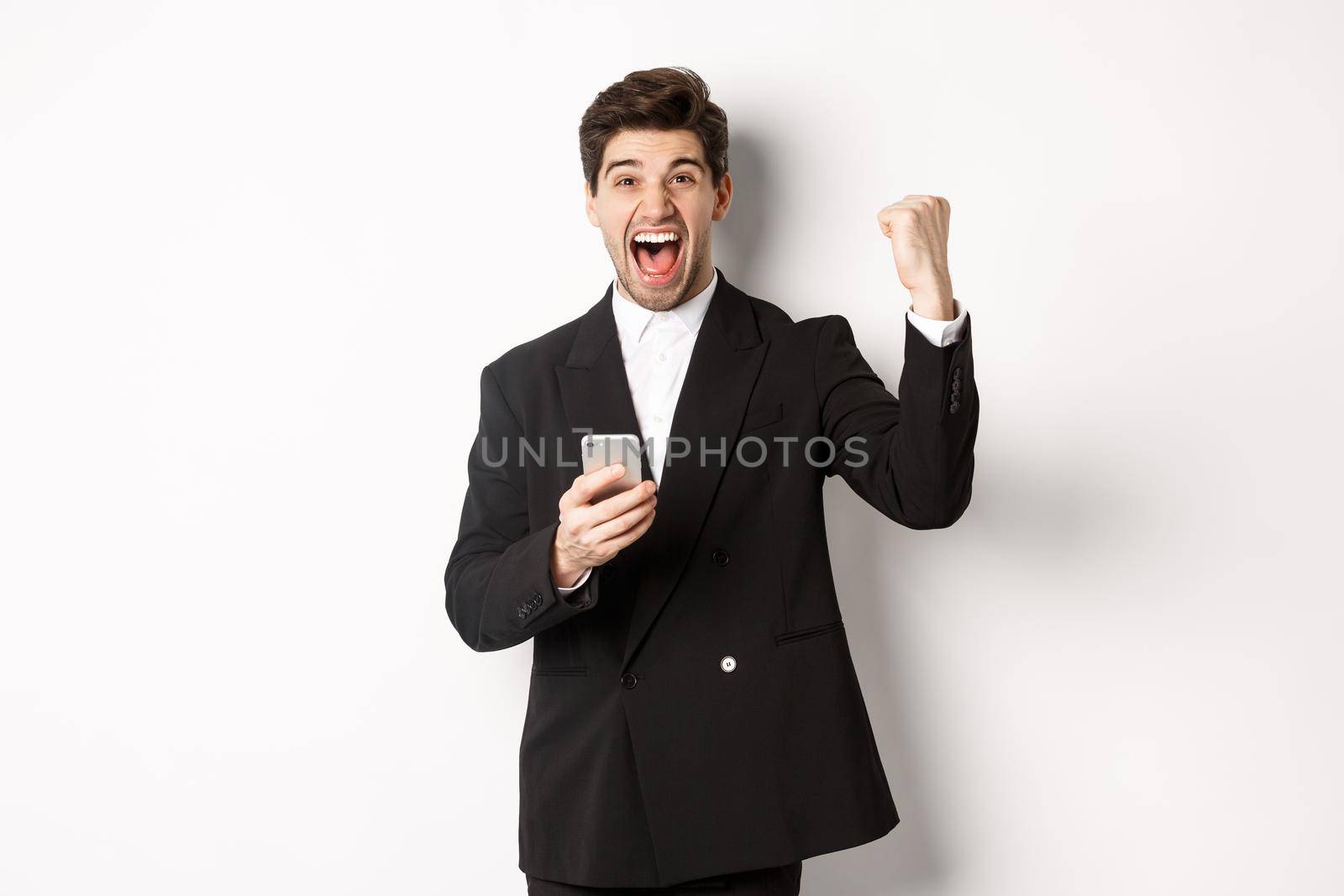 Portrait of happy handsome man in suit, rejoicing, achieve goal on mobile app, raising fist up and shouting yes, holding smartphone, standing against white background.