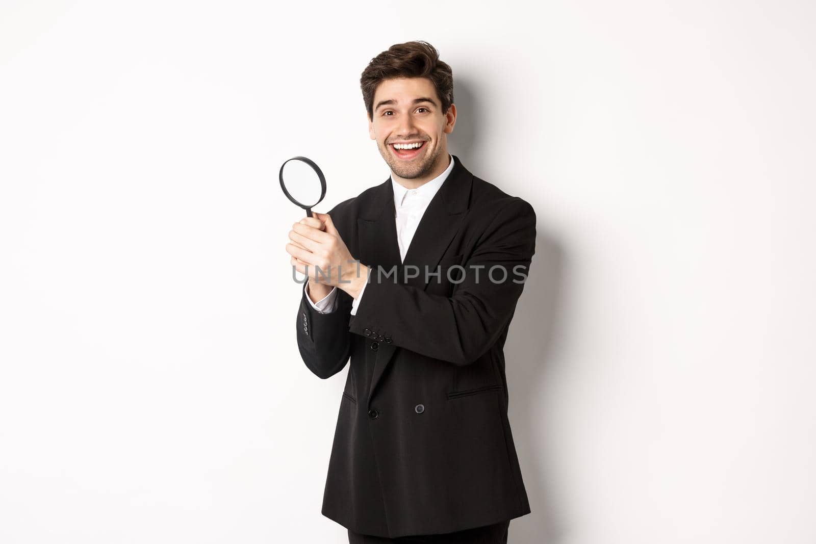 Handsome businessman in black suit, holding magnifying glass and smiling, looking for you, standing against white background by Benzoix