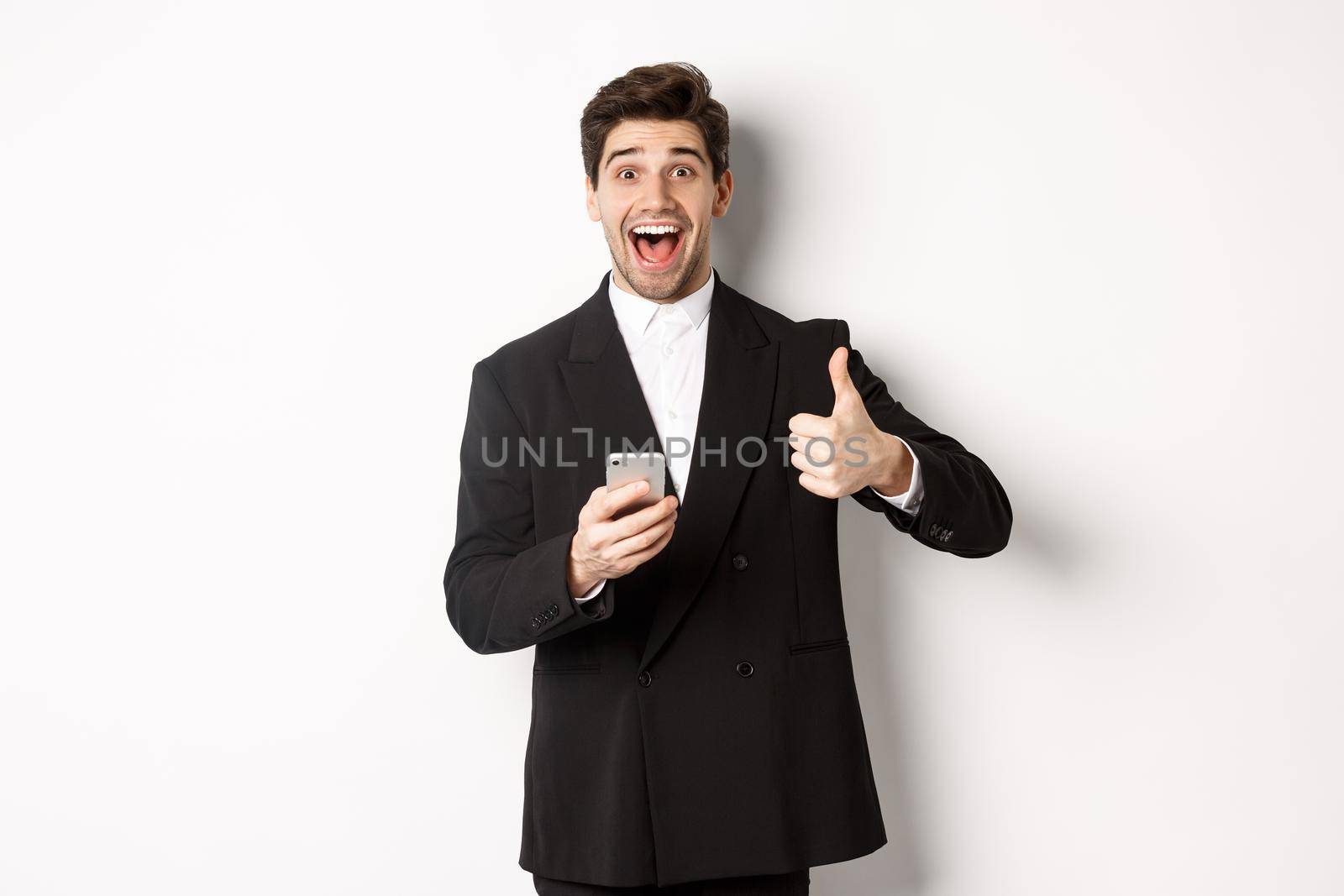 Portrait of handsome man in trendy suit showing thumbs-up in approval, using mobile phone app, smiling pleased, standing over white background.