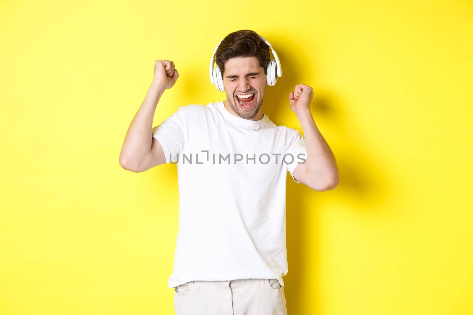 Happy man dancing and listening music in white headphones, standing over yellow background. Copy space