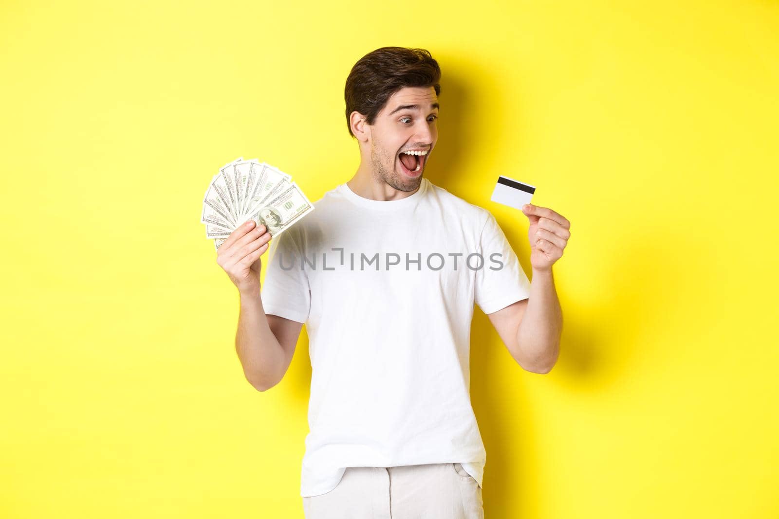 Cheerful guy looking at credit card, holding money, concept of bank credit and loans, standing over yellow background by Benzoix