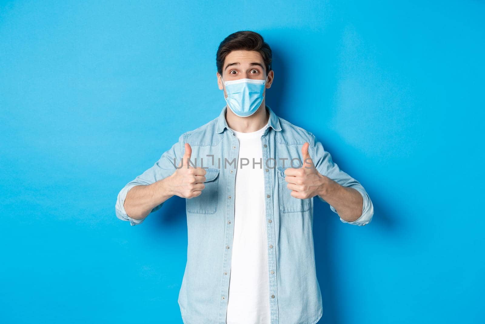 Concept of covid-19, pandemic and social distancing. Amazed guy in medical mask showing thumbs-up, recommending promo offer, standing against blue background.