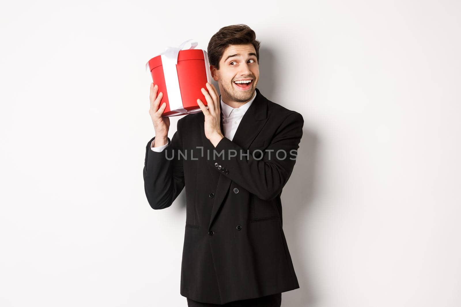Concept of christmas holidays, celebration and lifestyle. Handsome man in black suit, shaking a box with gift, wondering whats inside, standing against white background.