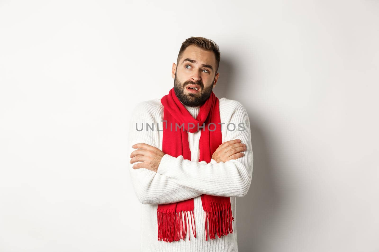 Confused man looking doubtful, standing with hands crossed and feeling cold on winter, wearing christmas sweater with red scarf, standing over white background by Benzoix