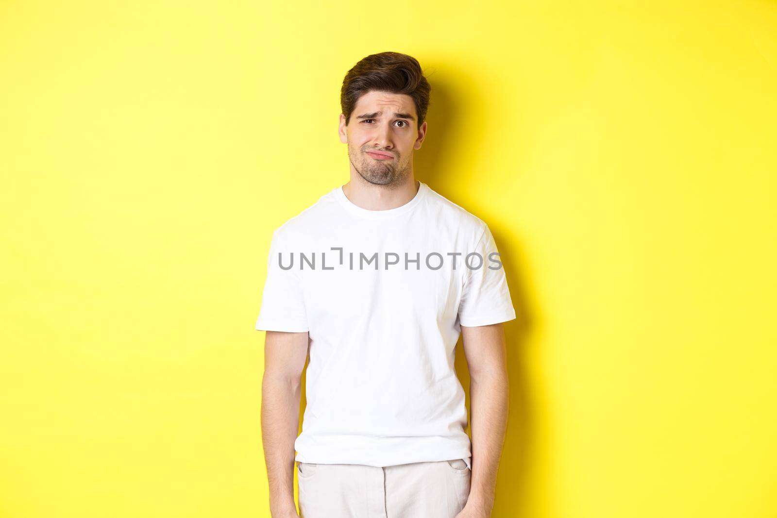 Disappointed guy looking upset, sulking and frowning, standing displeased against yellow background.
