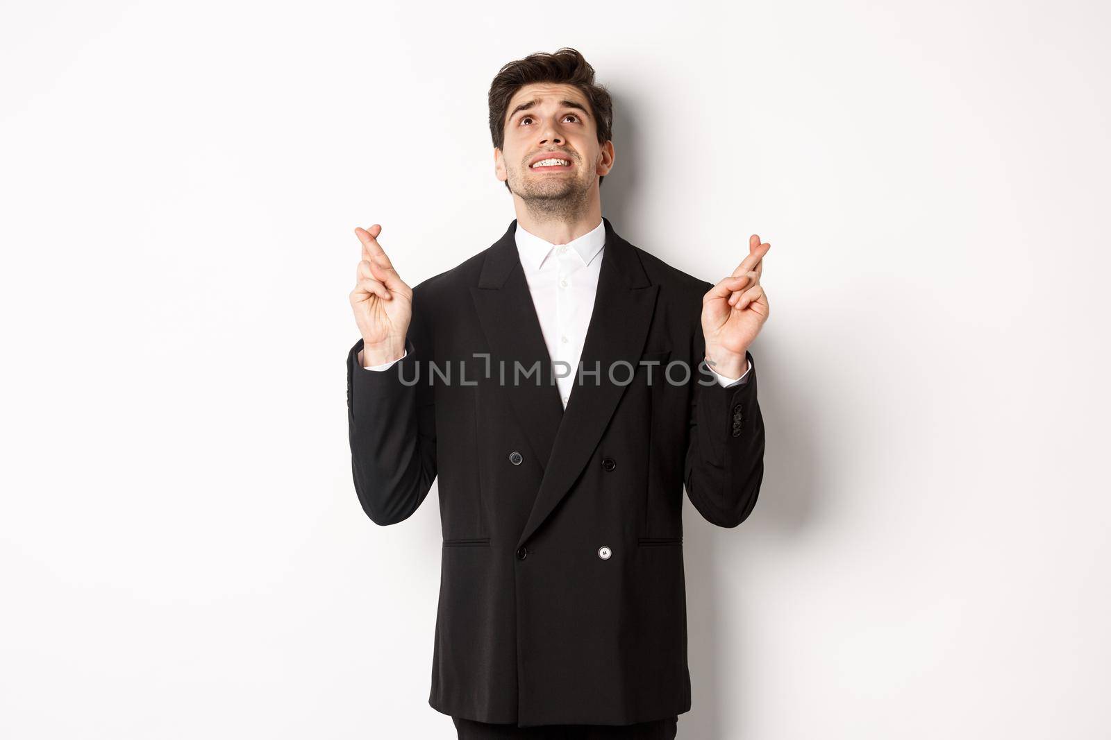 Portrait of tensed and worried handsome businessman, crossing fingers and looking up, begging god, making a wish, standing against white background in black suit by Benzoix
