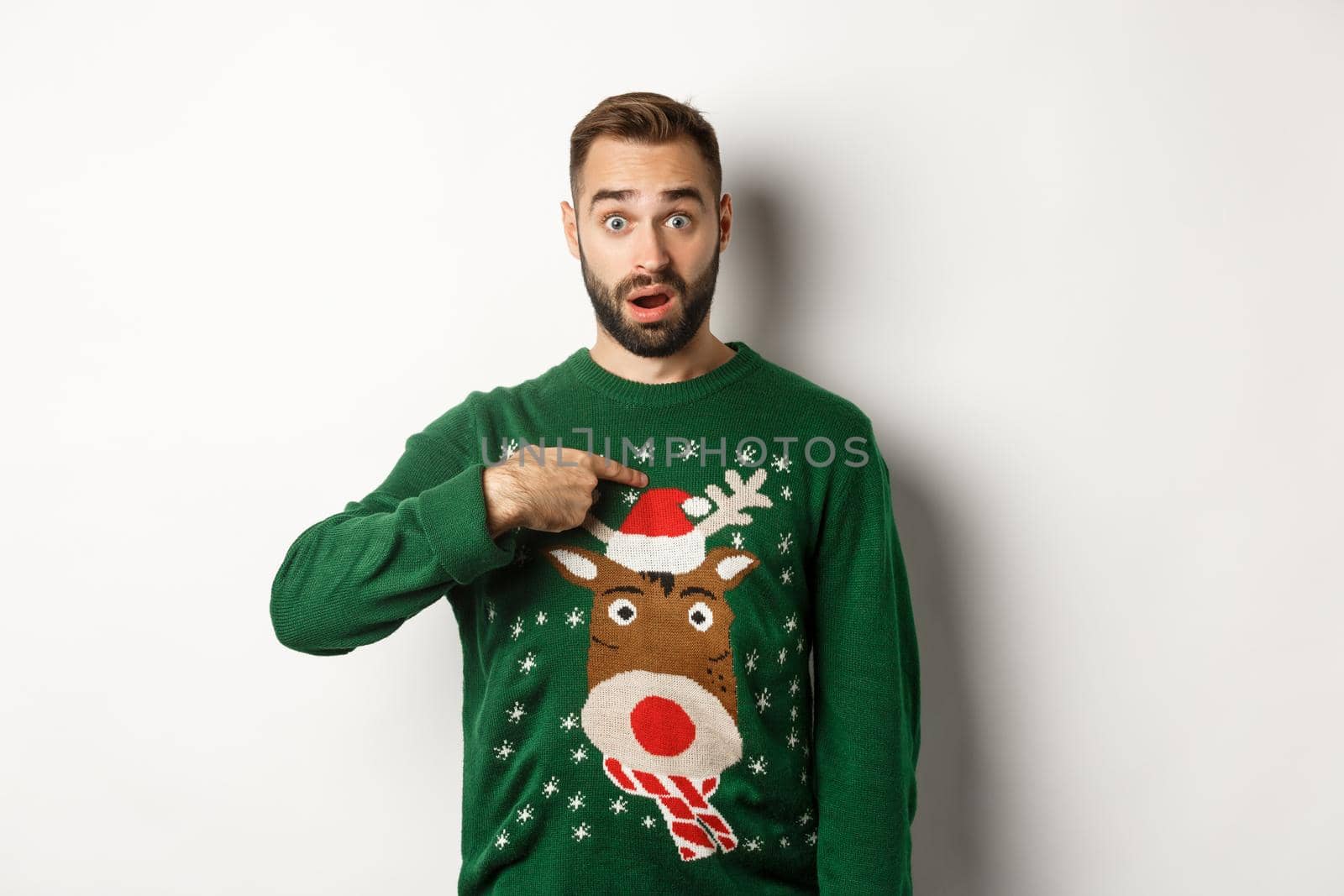 Winter holidays and christmas. Confused bearded guy pointing at himself, being startled with offer, standing over white background in sweater.