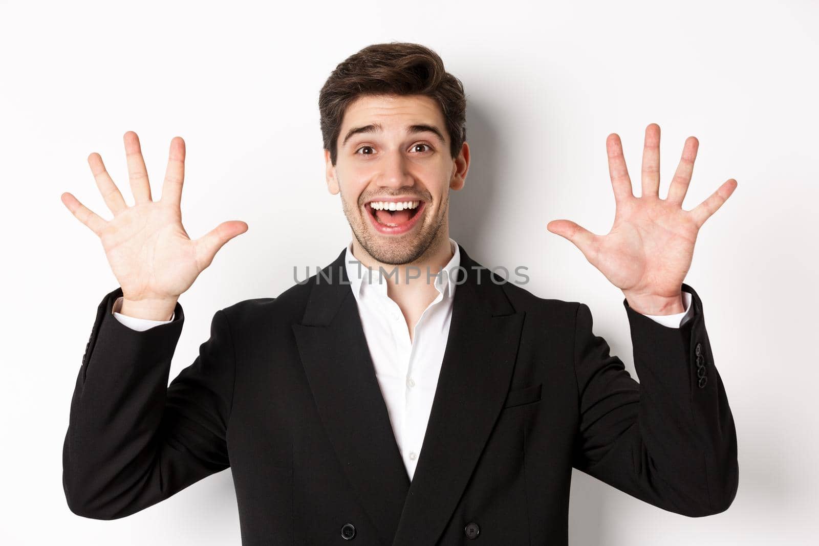 Close-up of handsome businessman in black suit, smiling amazed, showing number ten, standing over white background by Benzoix